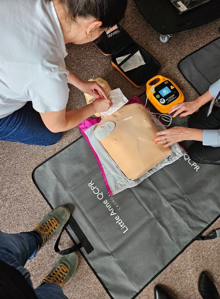 Kurs pierwszej pomocy: Instruktorzy ćwiczą resuscytację na manekinie Little Anne QCPR z użyciem defibrylatora AED Trainer. Widoczne notatki i akcesoria.