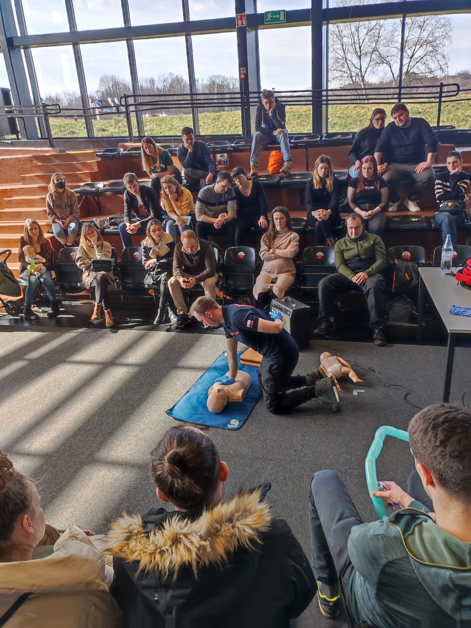 Instruktor demonstruje resuscytację krążeniowo-oddechową na manekinie podczas kursu pierwszej pomocy dla grupy osób w sali z dużymi oknami.