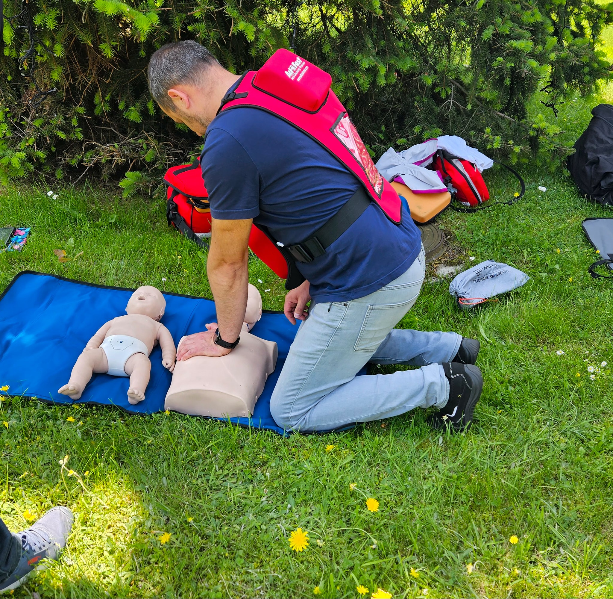 Instruktor demonstruje resuscytację krążeniowo-oddechową na manekinie na trawie, obok leży manekin niemowlęcia. Widoczny plecak ratunkowy.
