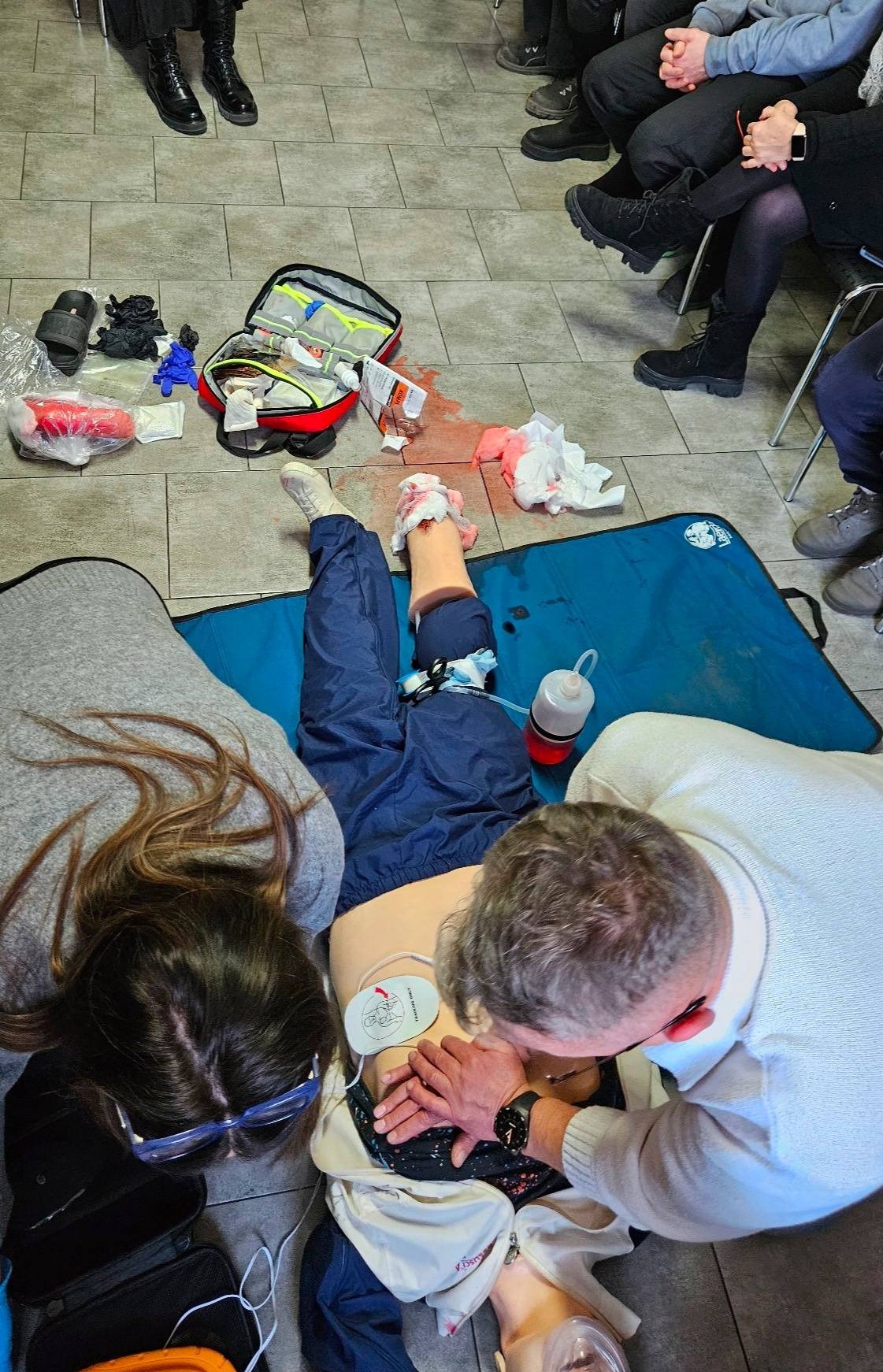 Kurs pierwszej pomocy: Manekin z raną nogi, otwarty zestaw ratunkowy, uczestnicy ćwiczą resuscytację krążeniowo-oddechową pod okiem instruktora. Widok z góry.