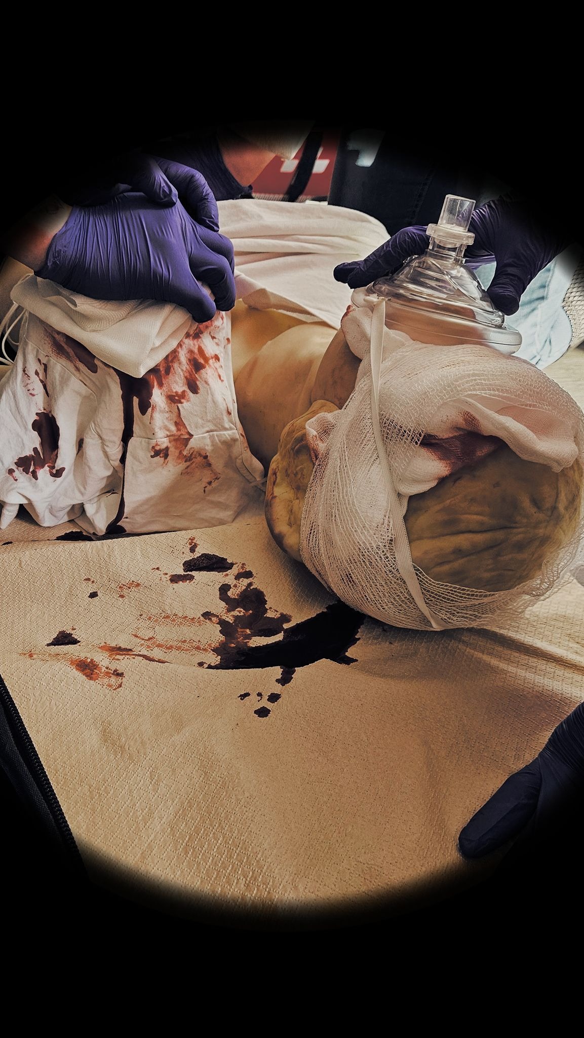 Symulacja udzielania pierwszej pomocy: manekin z bandażem na głowie i maską tlenową, krew na materiale, ręce w lateksowych rękawicach. Realistyczny trening medyczny.