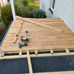 Budowa drewnianego tarasu: układanie desek na legarach, widoczna poziomica, wkrętarka i metalowe łączniki, dom w tle.