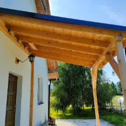 Drewniana altana przylegająca do białego budynku, z czarnym dachem, oświetlona girlandą żarówkową, widok na ogród i drzewa.