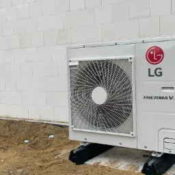 Biała zewnętrzna jednostka pompy ciepła LG Therma V, zamontowana na czarnych gumowych podstawach, na tle ściany z białych bloczków i fragmentu okna, z widoczną ziemią wokół.