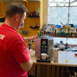 Serwisant w czerwonej koszulce z napisem 'KLIMATYZACJE SERWIS AGD' naprawia automat do kawy na stole warsztatowym, widoczne narzędzia i akcesoria.