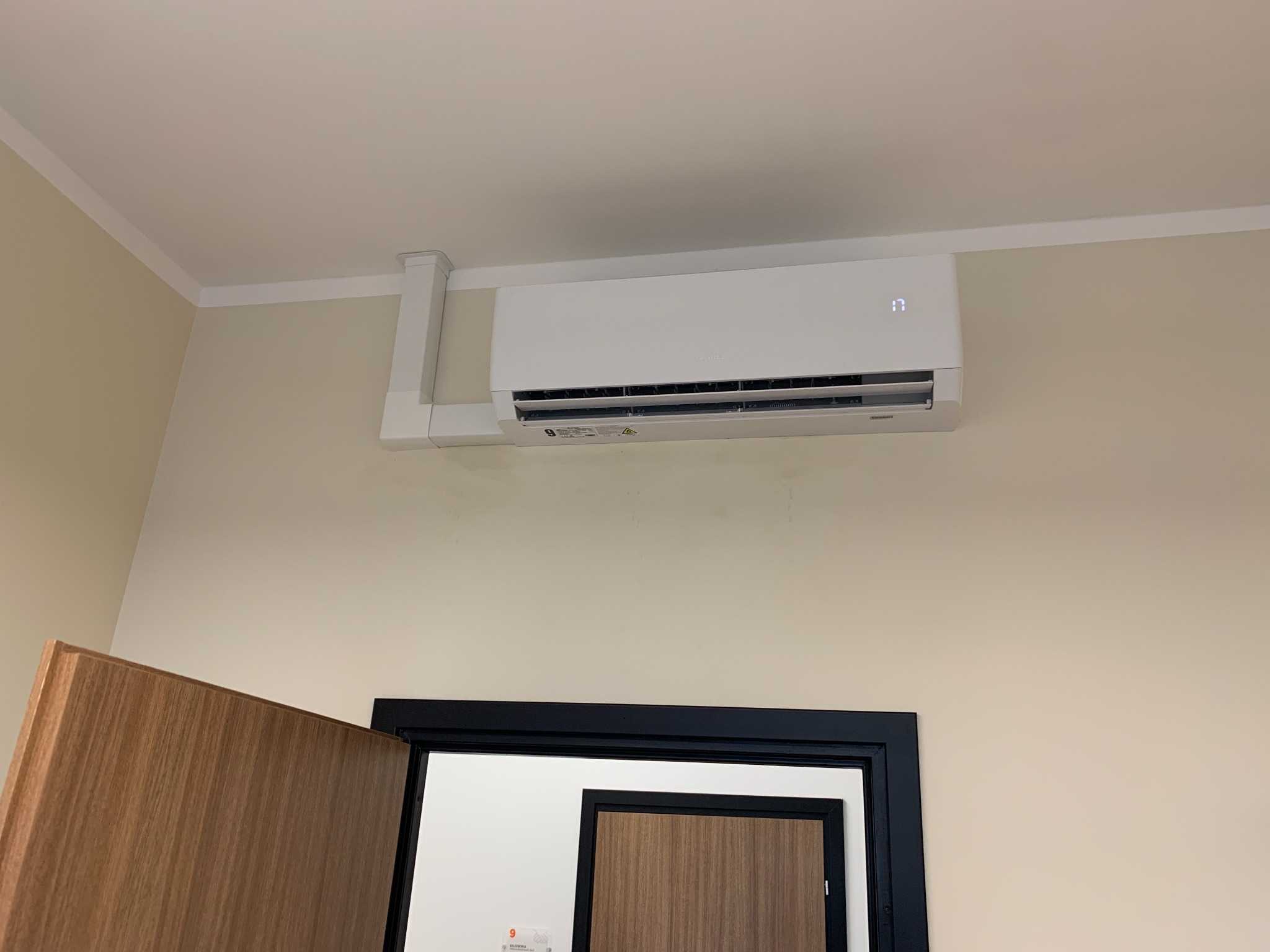 Zamontowany, biały klimatyzator GREE Pular z widoczną instalacją na kremowej ścianie, nad otwartymi drzwiami.