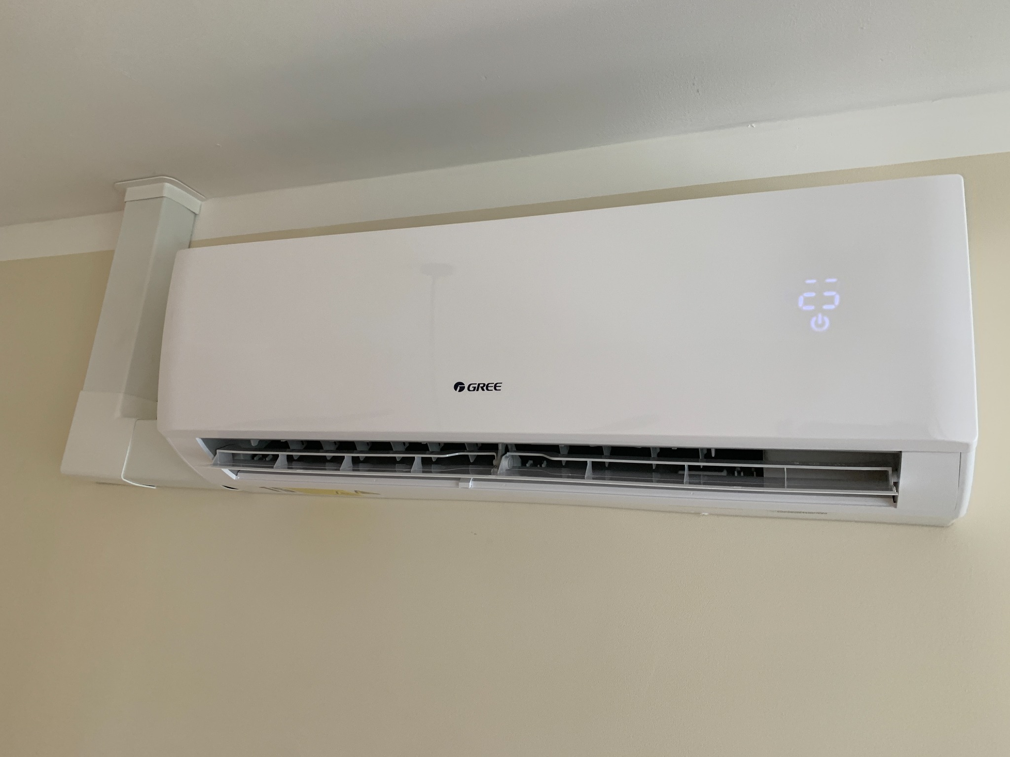 Biała klimatyzacja GREE Pular zamontowana na beżowej ścianie, widoczny wyświetlacz temperatury i kanał maskujący instalację.
