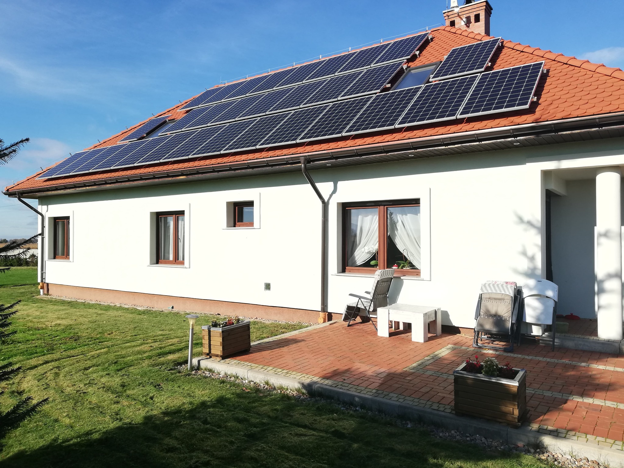 Dom jednorodzinny z panelami słonecznymi na dachu z czerwonej dachówki, widok z ogrodu w słoneczny dzień.