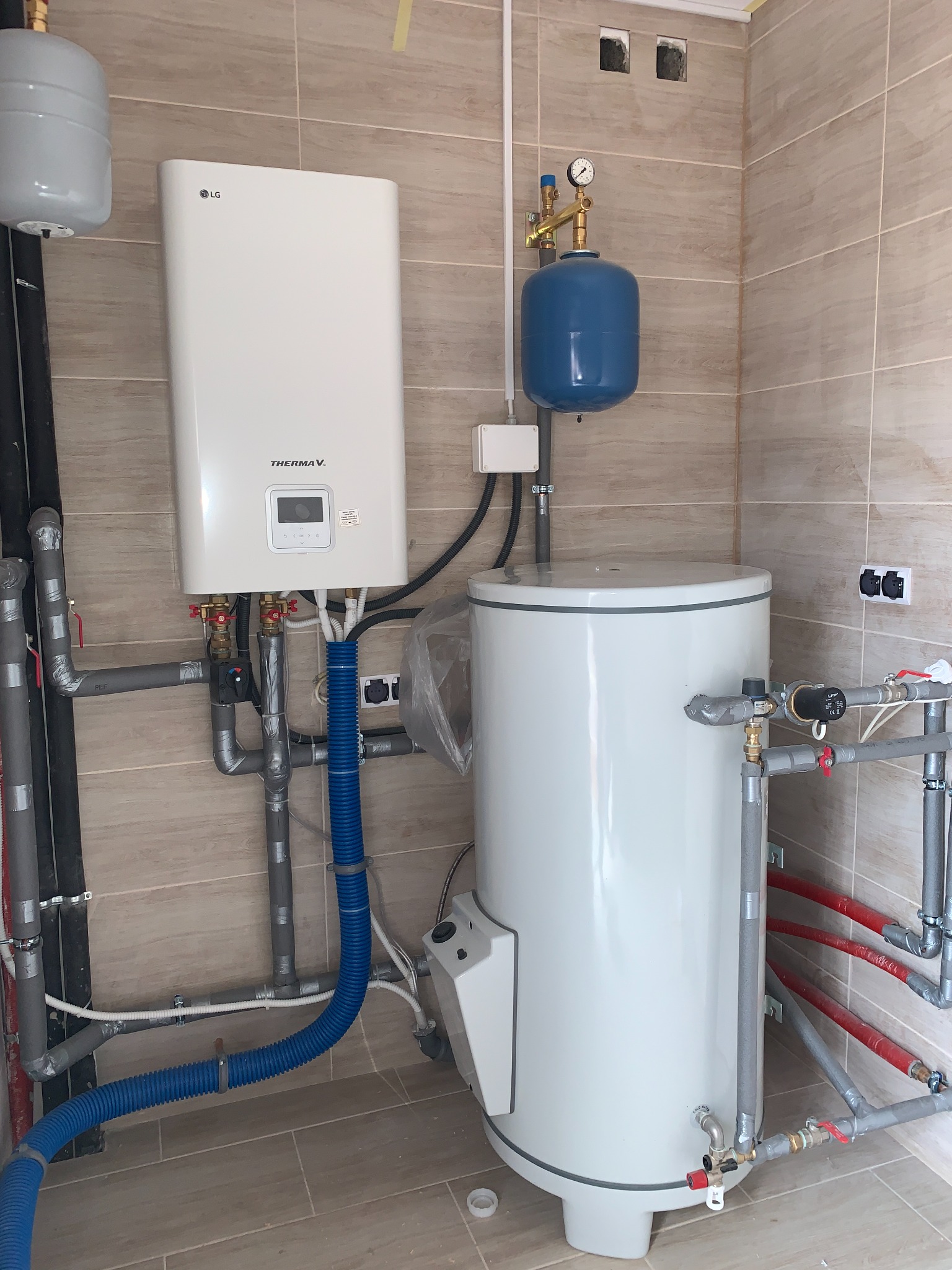 Instalacja kotła grzewczego LG Therma V z zasobnikiem ciepłej wody w pomieszczeniu z beżowymi płytkami ściennymi i podłogowymi, widoczne liczne rury i zawory.