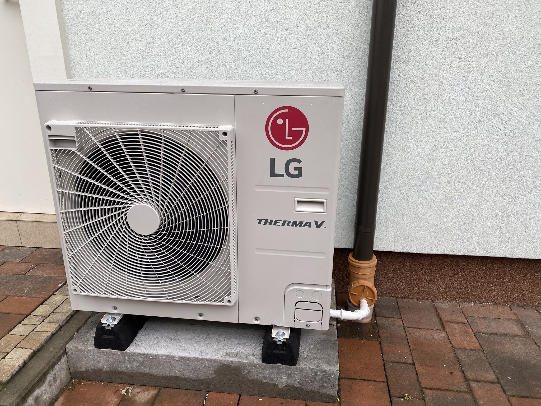 Pompa ciepła LG Therma V zamontowana na betonowym fundamencie z czarnymi podkładkami antywibracyjnymi, widoczny odpływ kondensatu i rynna spustowa przy ścianie budynku.
