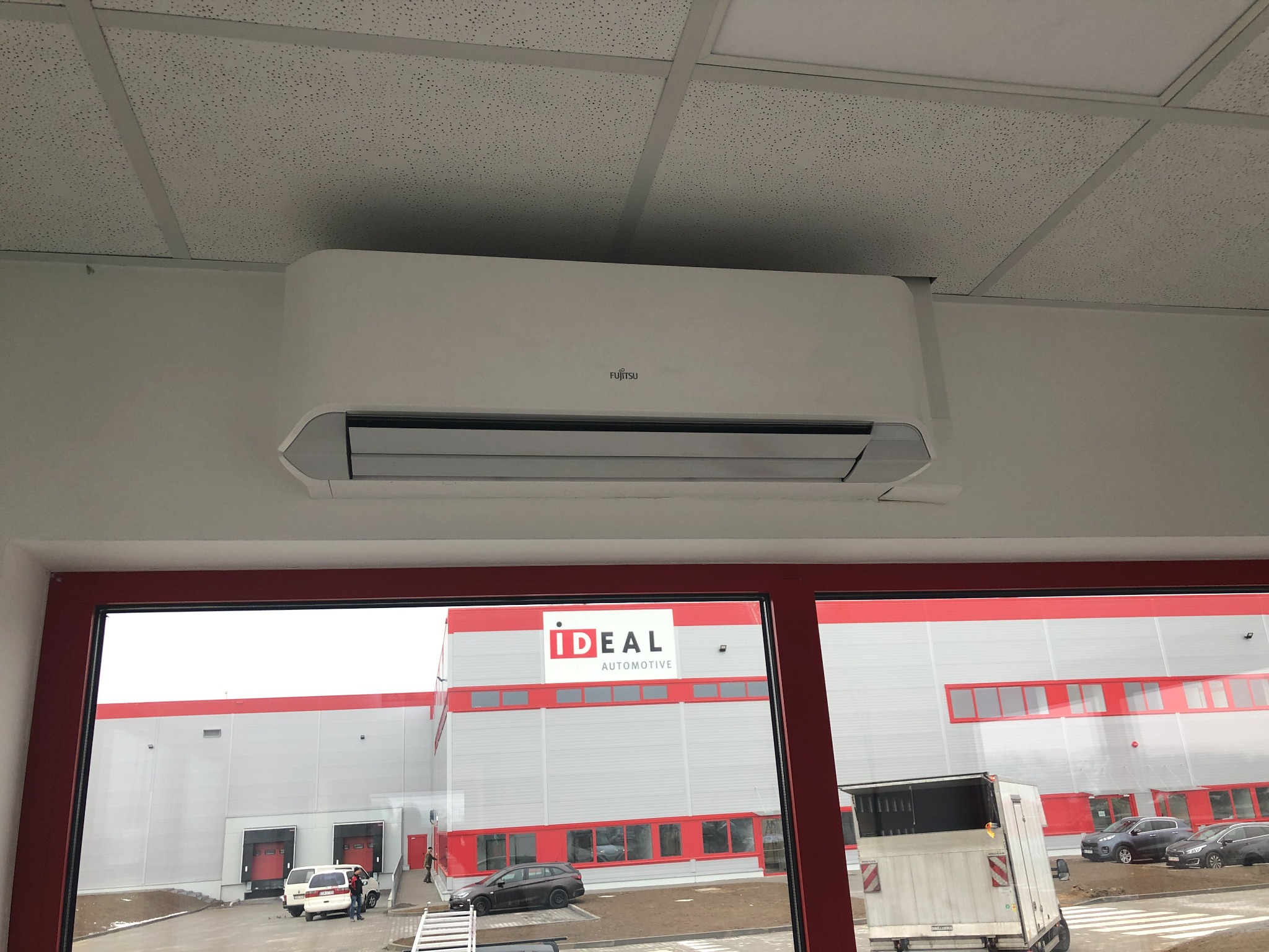 Biały klimatyzator Fujitsu zamontowany na ścianie w pomieszczeniu biurowym, widok z okna na budynek magazynowy z logo IDEAL AUTOMOTIVE.