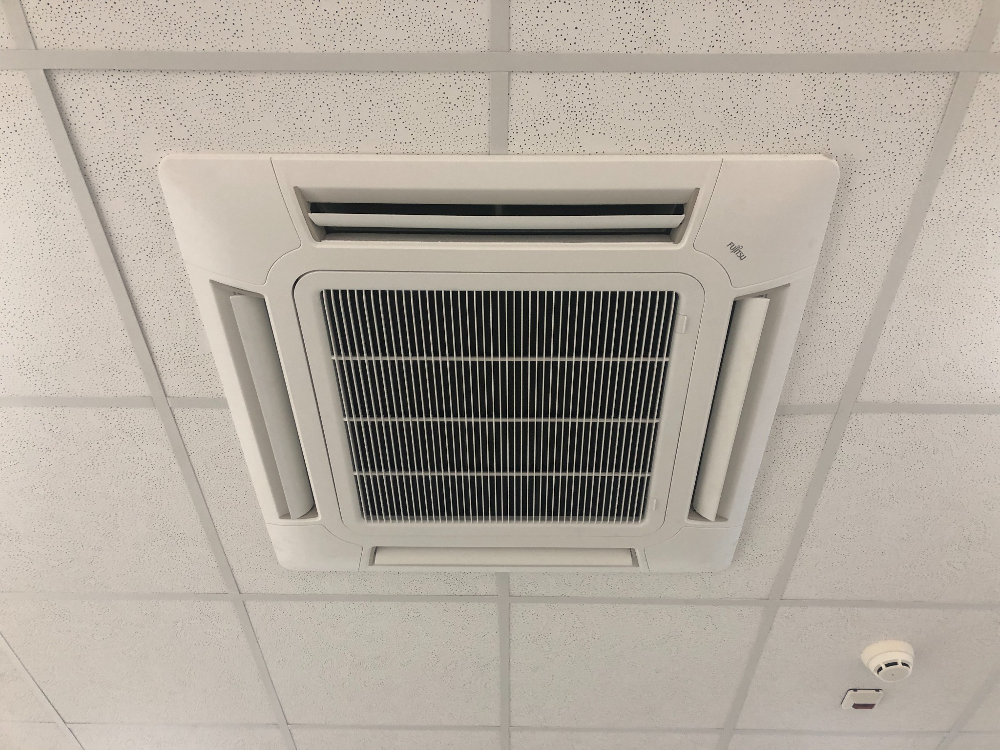Biały klimatyzator kasetonowy marki Fujitsu zamontowany w suficie podwieszanym z widocznym czujnikiem dymu w rogu.