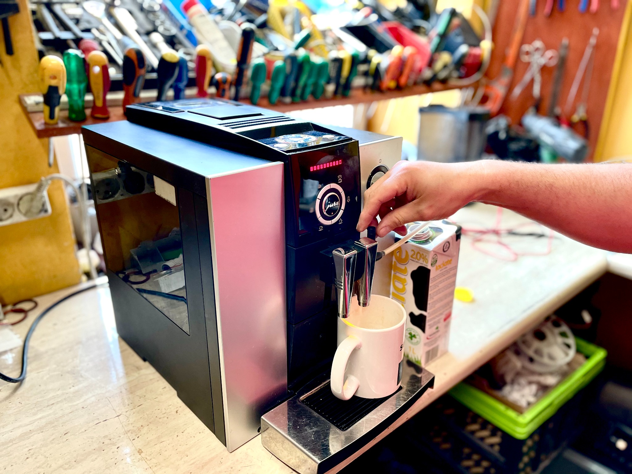 Ręka obsługuje automatyczny ekspres do kawy Jura, na tle ściany z narzędziami, obok karton mleka 2.0%.