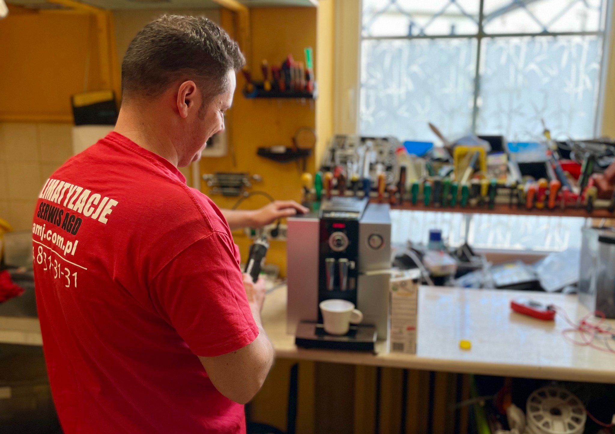 Serwisant w czerwonej koszulce z napisem 'KLIMATYZACJE SERWIS AGD' naprawia automat do kawy na stole warsztatowym, widoczne narzędzia i akcesoria.