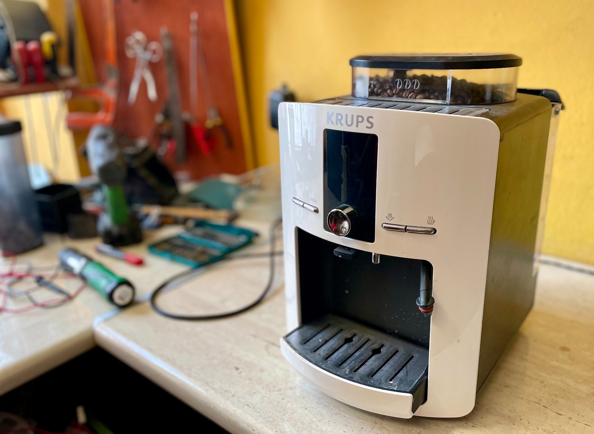 Ekspres do kawy Krups z widocznymi ziarnami kawy w pojemniku, stojący na blacie warsztatowym z narzędziami w tle.