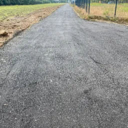 Nowo położona, czarna nawierzchnia asfaltowa drogi dojazdowej, widok z perspektywy pieszego, z widocznym polem po lewej stronie i ogrodzeniem z siatki po prawej, w tle zabudowania.