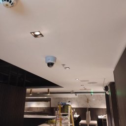 Usługi Elektroinstalacyjne Rafał Tokarz - Sufit w lokalu komercyjnym z zamontowanymi czujnikami dymu, kamerą przemysłową kopułkową oraz oświetleniem punktowym; żółta drabina rozstawiona w tle, sugerująca trwające prace instalacyjne.