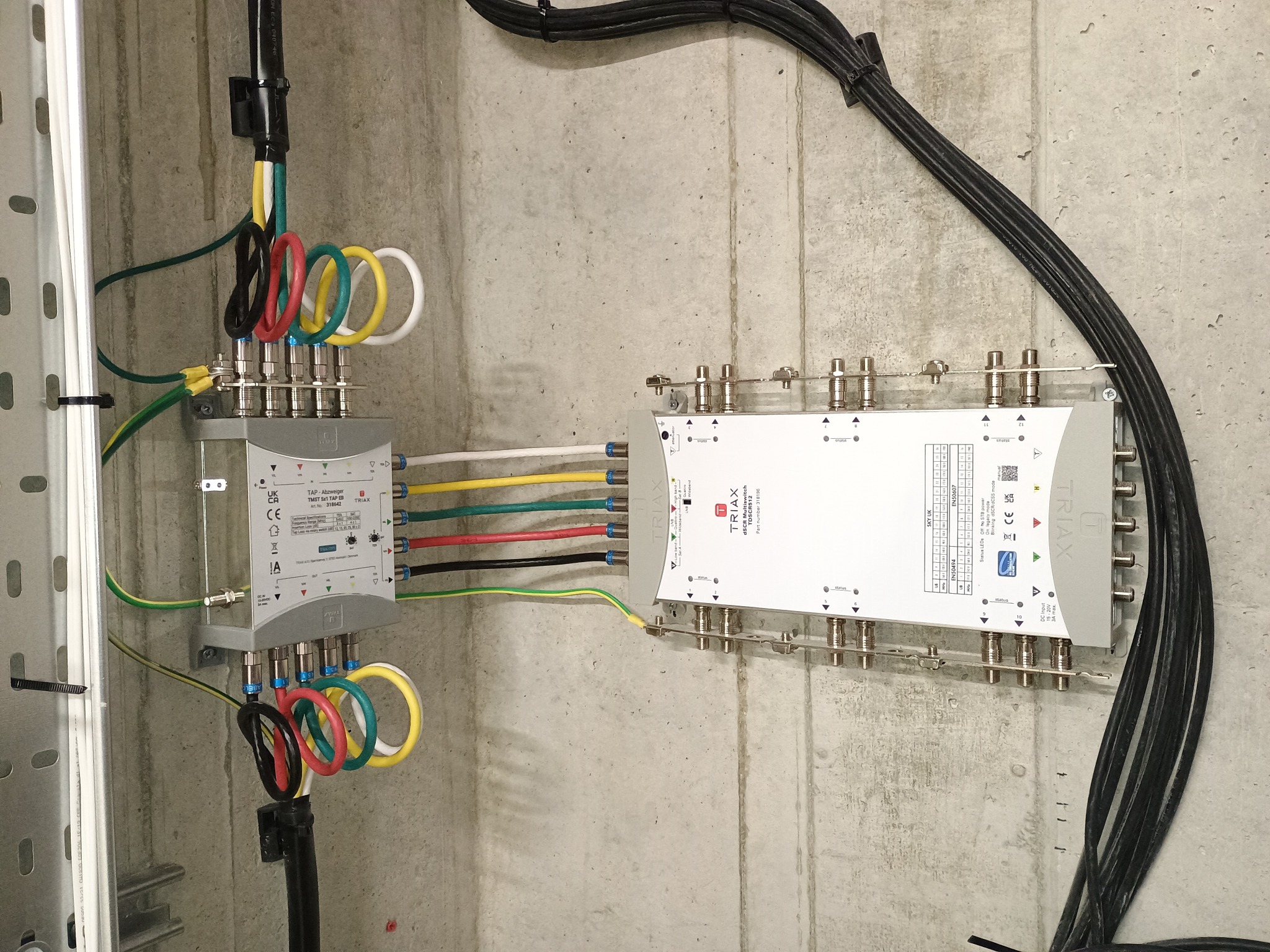 Dwa rozdzielacze sygnału TV marki Triax zamontowane na betonowej ścianie, z podłączonymi kablami koncentrycznymi w kolorowych pętlach oraz czarnymi kablami w wiązce. Widoczna szyna montażowa po...