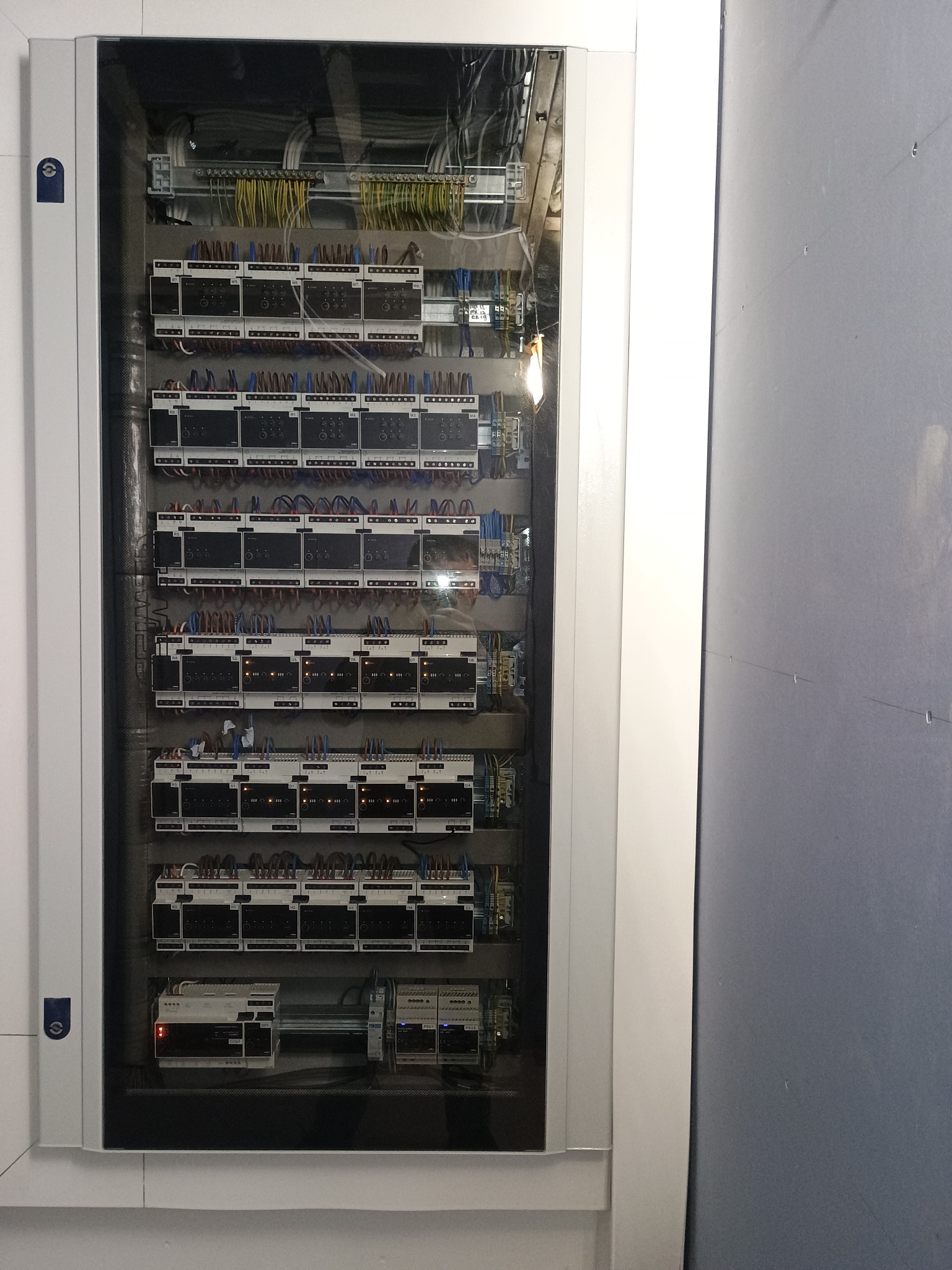 Rozdzielnica elektryczna w białej obudowie, widoczne liczne moduły sterujące z okablowaniem, zamontowane w szafie na szarej ścianie.