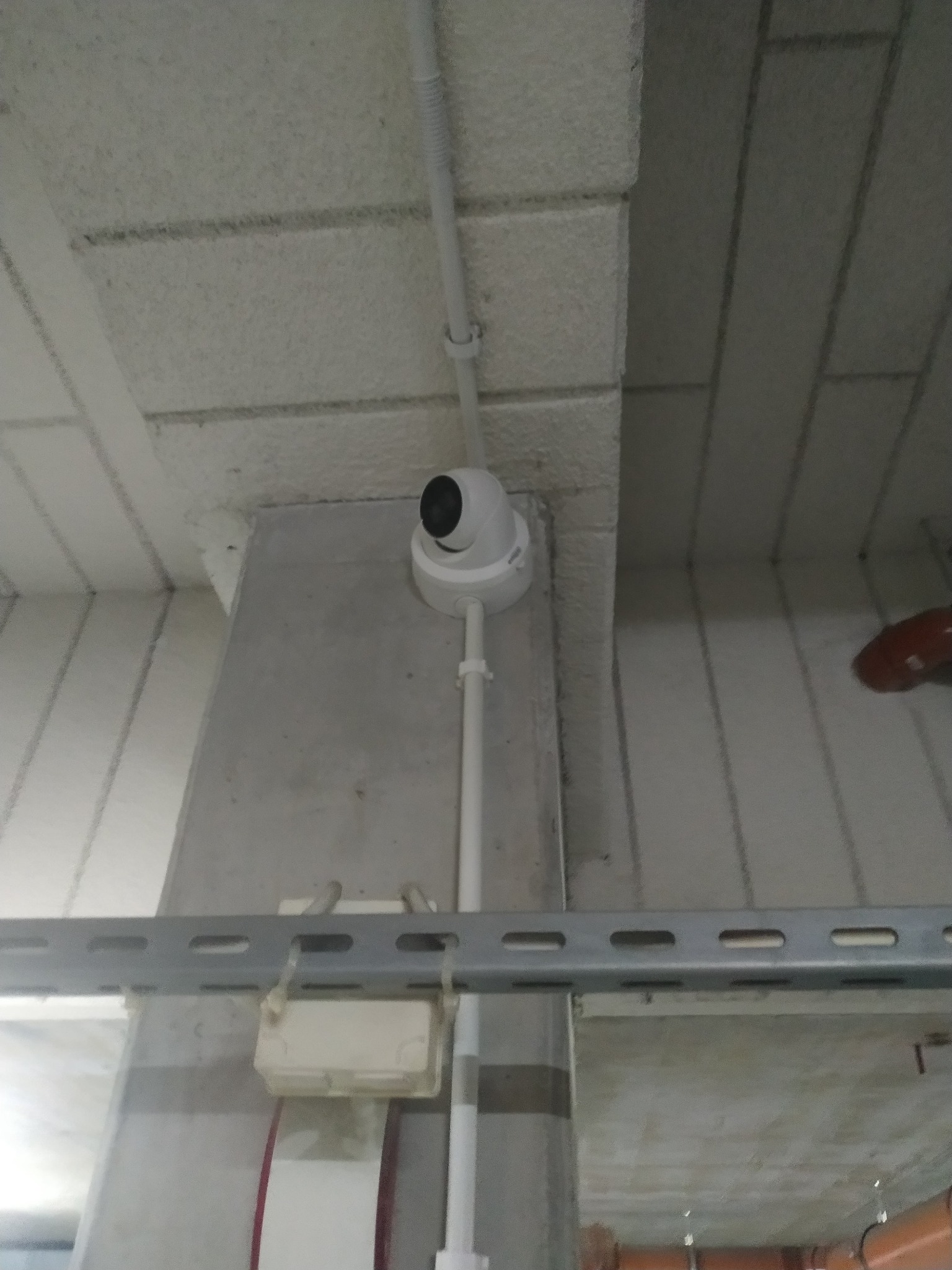 Kamera monitoringu kopułkowa zamontowana na białej rurze pionowej, widoczna pod sufitem w pomieszczeniu przemysłowym, z szarą belką perforowaną w tle.