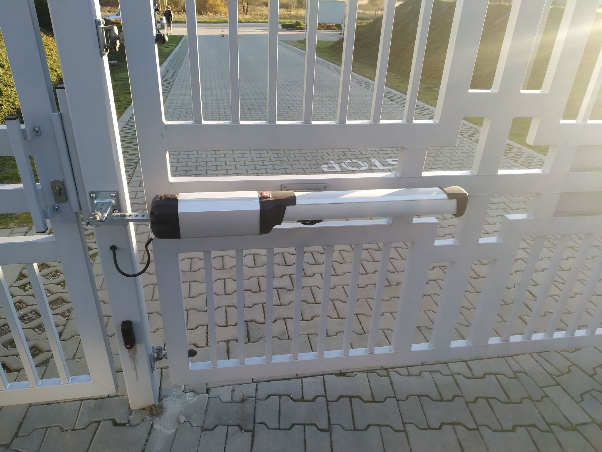 Biała, metalowa brama wjazdowa z zamontowanym siłownikiem automatycznym, widoczny czytnik kart dostępu na słupku, kostka brukowa na podjeździe.