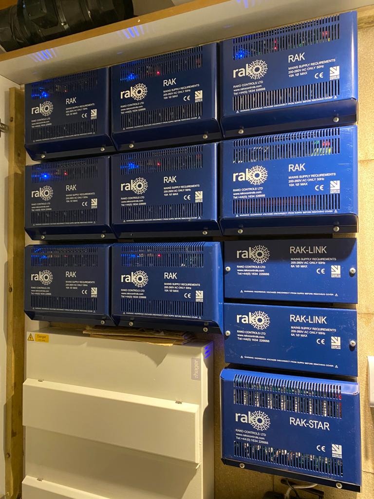 Ściana z zamontowanymi niebieskimi zasilaczami RAK różnych modeli (RAK, RAK-LINK, RAK-STAR) i białą skrzynką rozdzielczą Hager pod spodem.