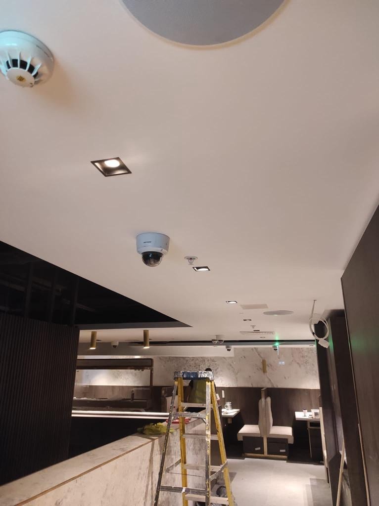 Sufit w lokalu komercyjnym z zamontowanymi czujnikami dymu, kamerą przemysłową kopułkową oraz oświetleniem punktowym; żółta drabina rozstawiona w tle, sugerująca trwające prace instalacyjne.