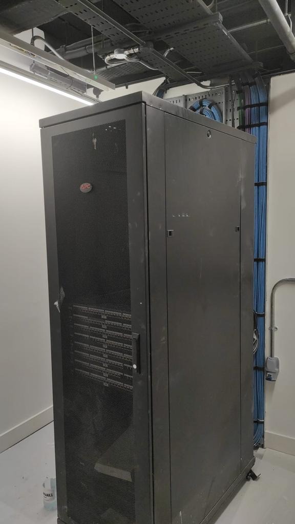 Czarna szafa serwerowa APC z widocznymi kablami sieciowymi w tle, umieszczona w pomieszczeniu z surowym wykończeniem, oświetleniem LED i instalacjami pod sufitem.