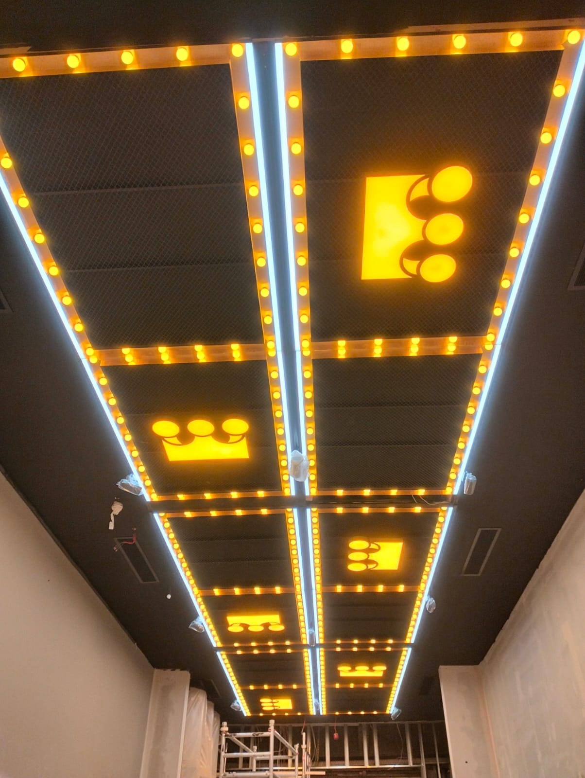 Sufit z podwieszanymi panelami oświetleniowymi w kształcie koron i neonami, widoczne elementy instalacji elektrycznej i rusztowanie.