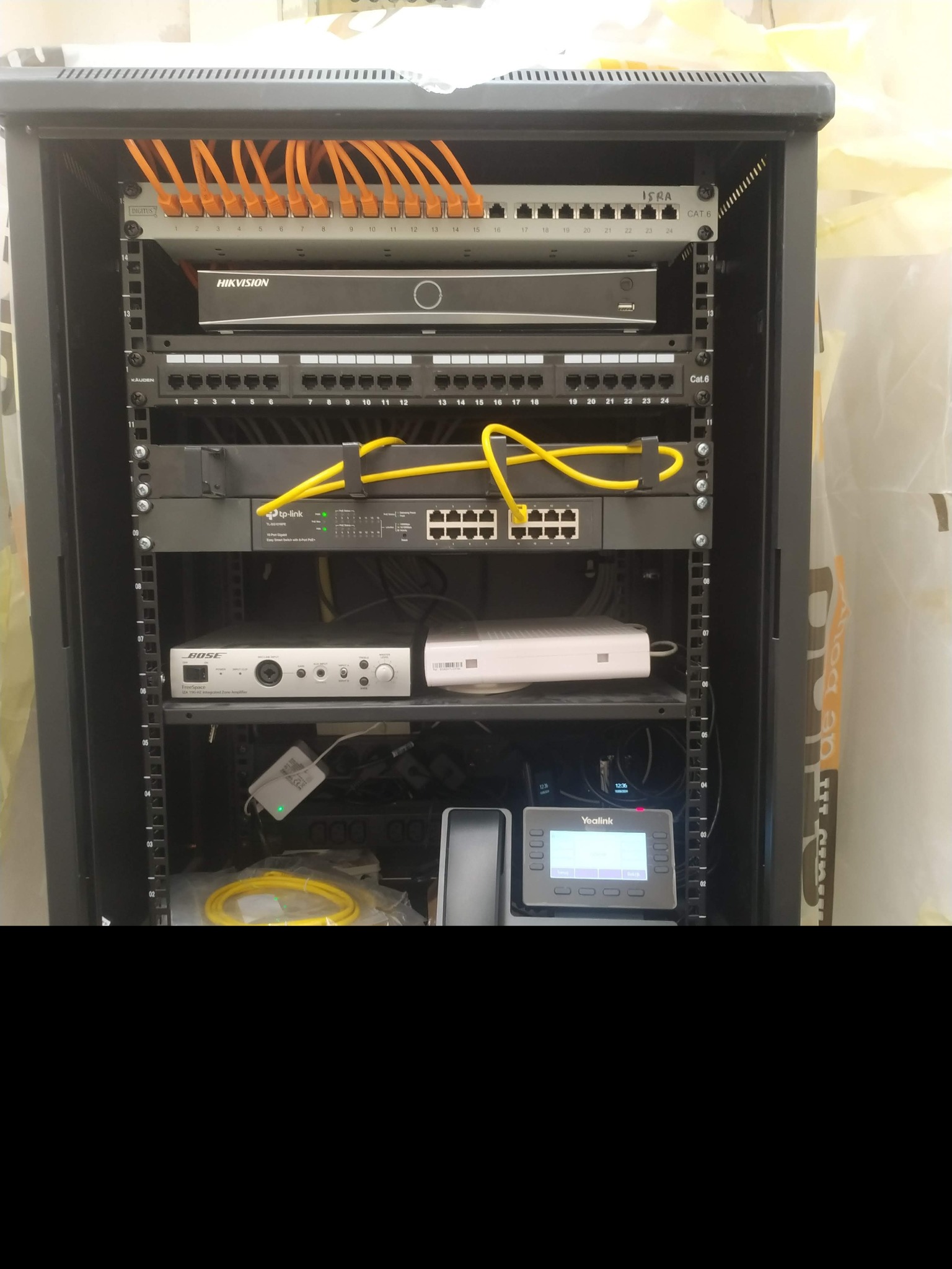 Szafa rack z zamontowanym rejestratorem Hikvision, switchem TP-Link, wzmacniaczem Bose i telefonem Yealink, okablowanie strukturalne z pomarańczowymi kablami patchcord. Widoczne złącza Cat.6.
