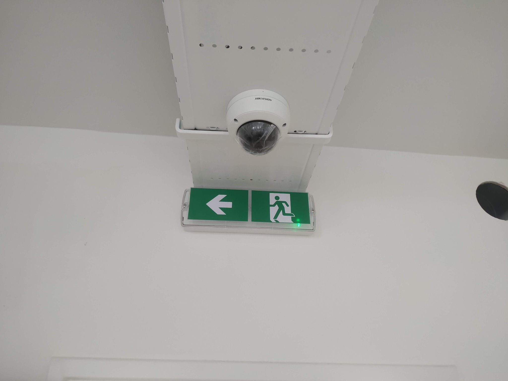 Kopułkowa kamera monitoringu Hikvision zamontowana pod sufitem, obok podświetlonego znaku ewakuacyjnego ze strzałką w lewo.