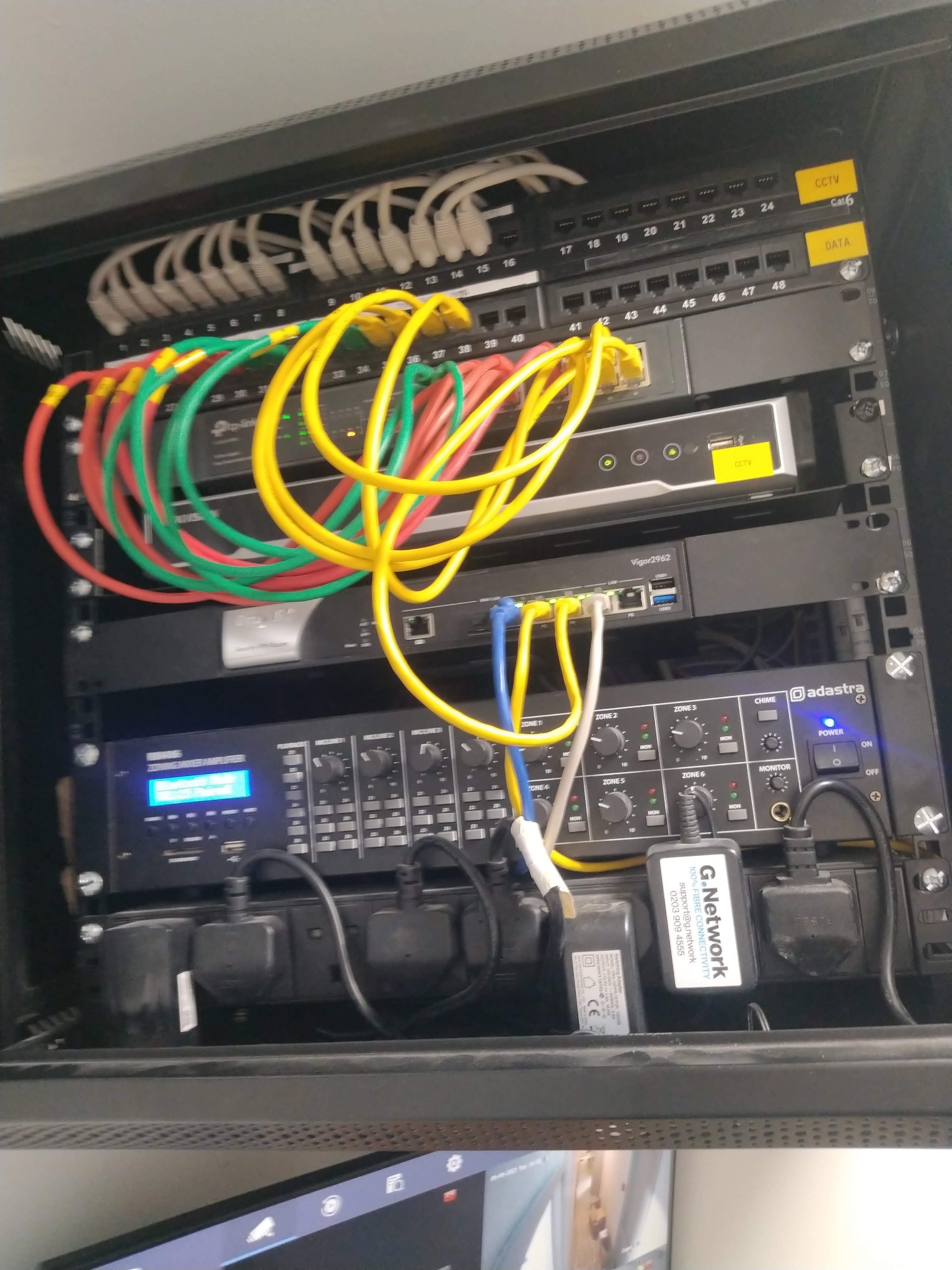 Szafa rack z zainstalowanymi urządzeniami sieciowymi, patch panelem, routerem, wzmacniaczem audio i zasilaczami, okablowanie w kolorach czerwonym, zielonym i żółtym, etykiety CCTV i DATA na patch...