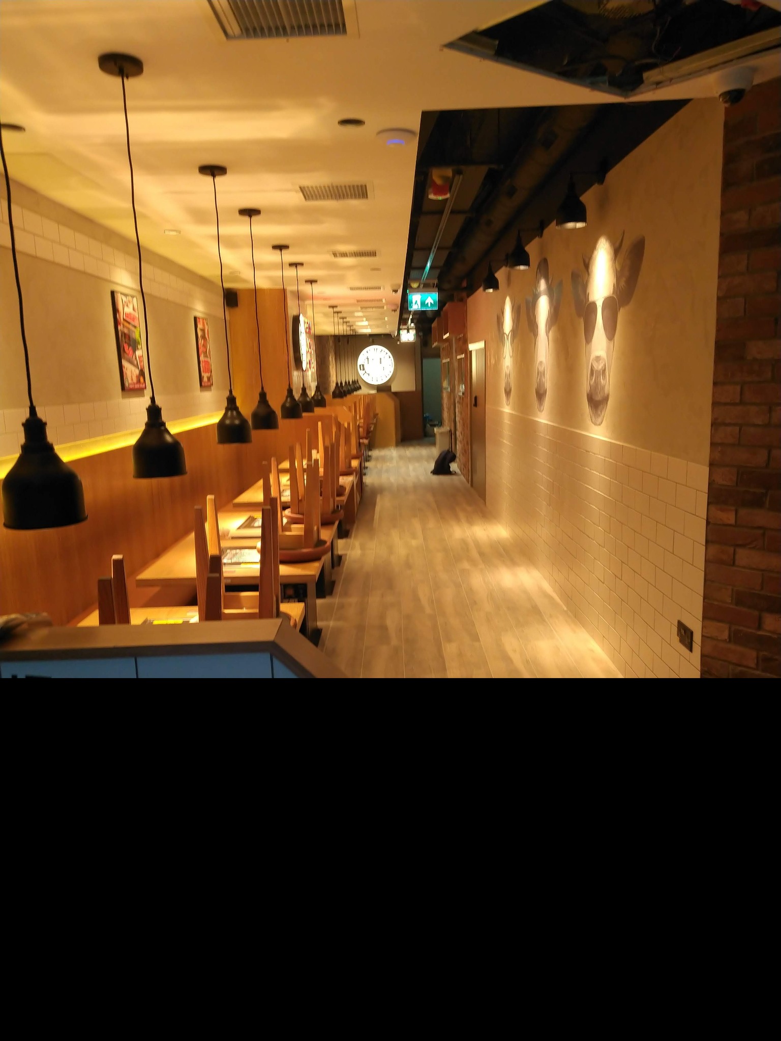 Wnętrze restauracji z rzędem stolików i krzeseł ustawionych na nich, wiszące lampy w stylu industrialnym, na ścianach grafiki krów w okularach, widoczny fragment instalacji pod sufitem.