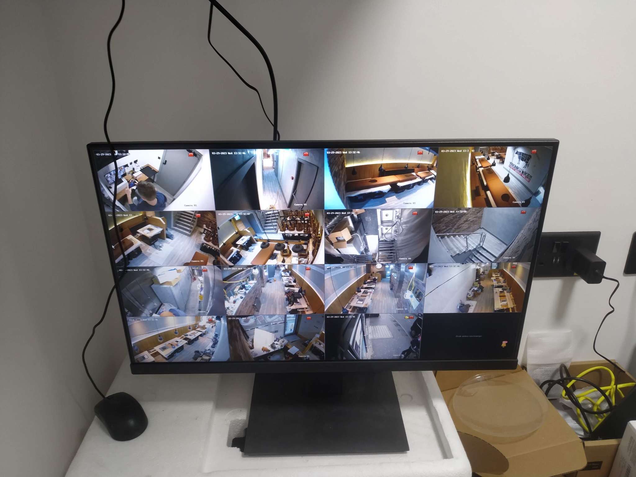 Monitor wyświetlający podgląd z wielu kamer systemu monitoringu wizyjnego, widoczne różne ujęcia wnętrz i otoczenia, obok mysz komputerowa i kable zasilające.