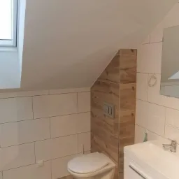 Nowoczesna łazienka na poddaszu ze skośnym sufitem, wykończona białymi i drewnopodobnymi płytkami, wisząca toaleta i biała umywalka z szafką, widok na okno dachowe.