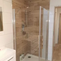 Nowoczesna kabina prysznicowa z przeszklonymi drzwiami, wyłożona płytkami imitującymi drewno i białymi płytkami, z widoczną armaturą i umywalką z szafką w tle.