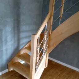 Jasne, drewniane schody z metalowymi prętami balustrady, widoczne z dołu, na tle szarej ściany i drewnianej podłogi.