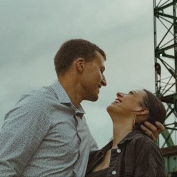 Agnieszka Górak - Uśmiechnięta para, mężczyzna obejmuje kobietę, spoglądają na siebie na tle industrialnego dźwigu i pochmurnego nieba.