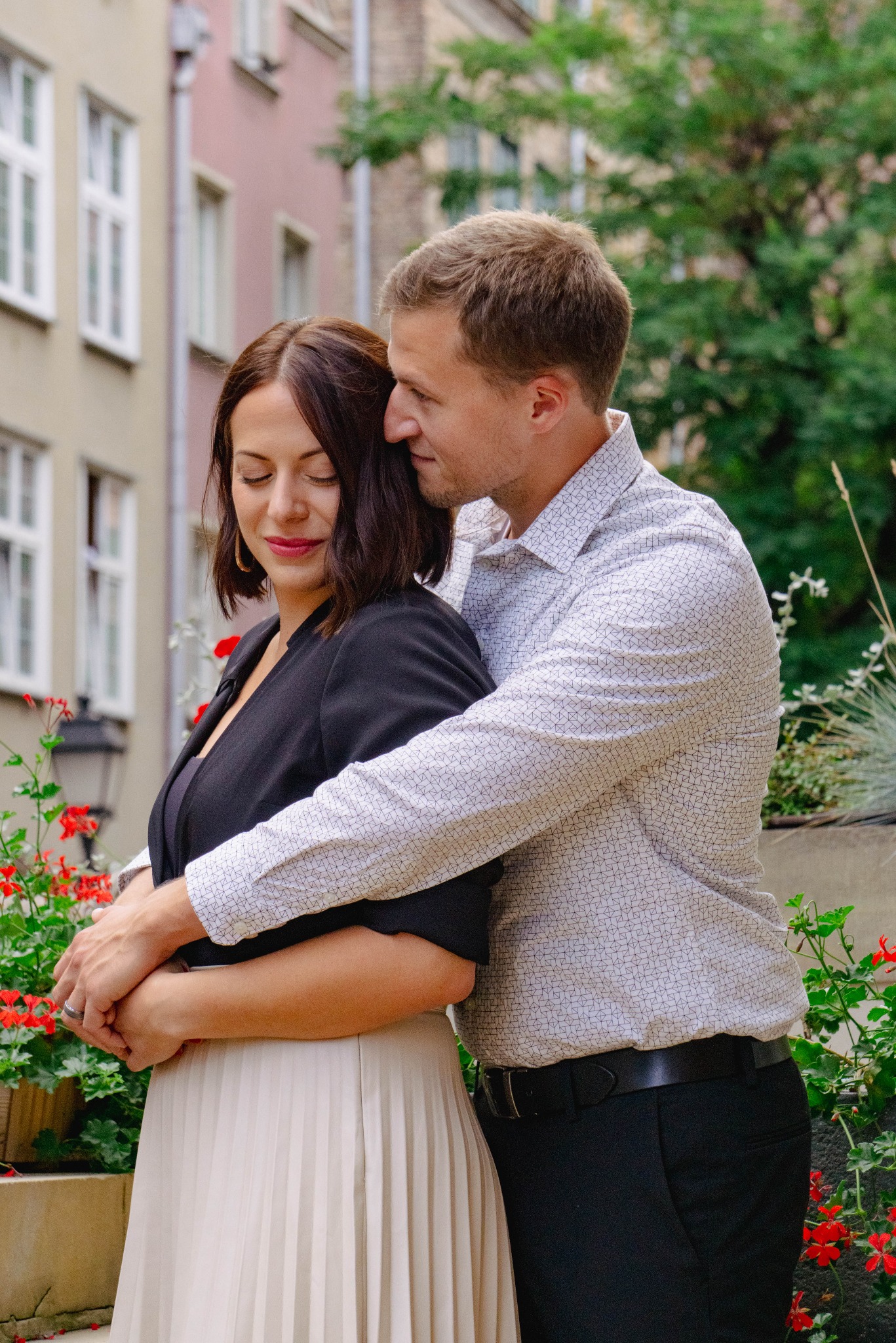 Para zakochanych: mężczyzna obejmuje kobietę w czarnej marynarce i plisowanej spódnicy na tle miejskiej zabudowy i czerwonych kwiatów.