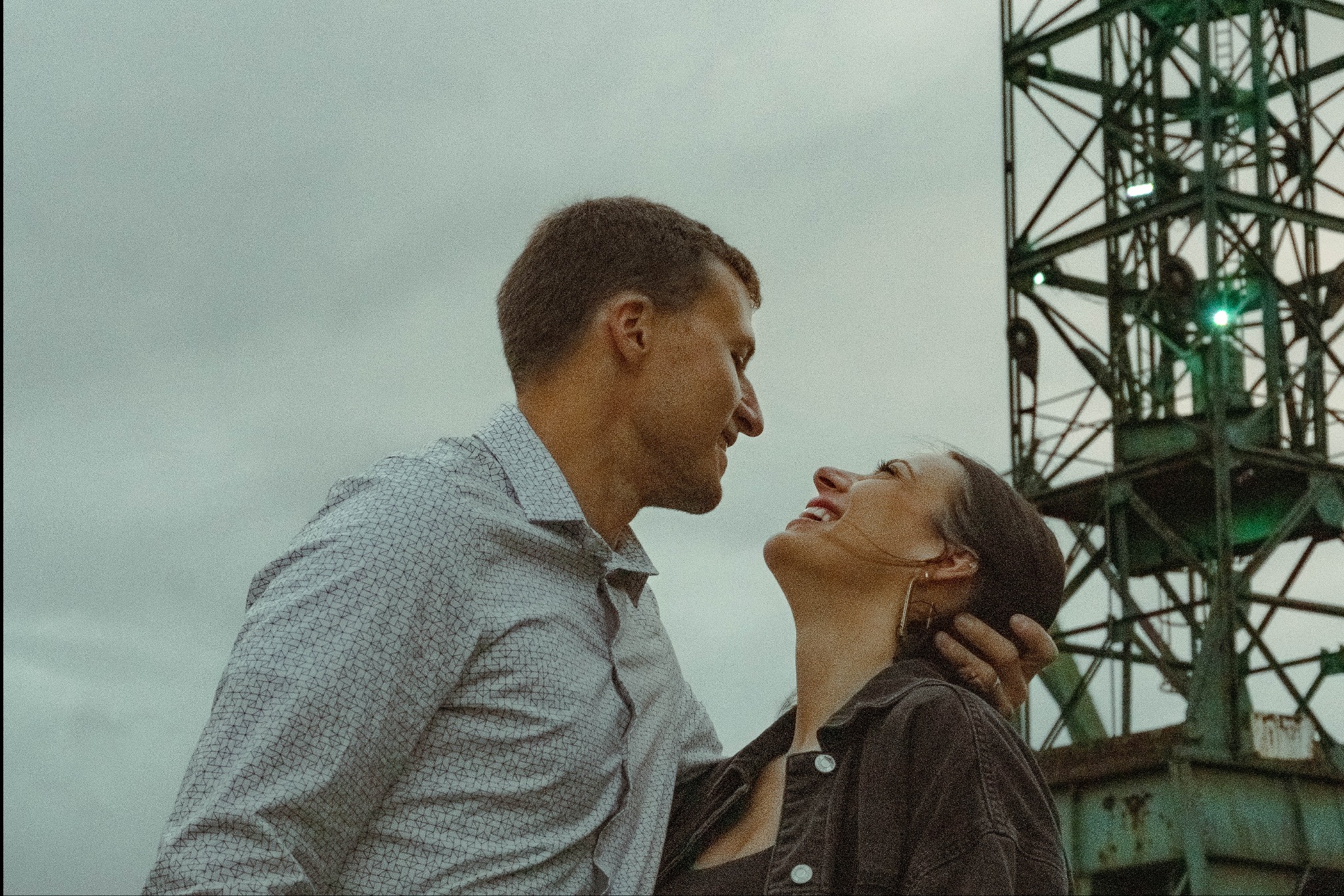 Uśmiechnięta para, mężczyzna obejmuje kobietę, spoglądają na siebie na tle industrialnego dźwigu i pochmurnego nieba.