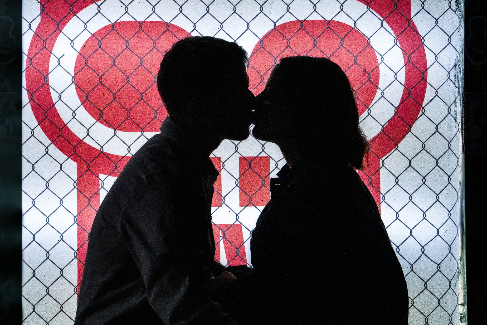 Silhouettes of a kissing couple against a backdrop of a chain-link fence and a red and white graphic design.