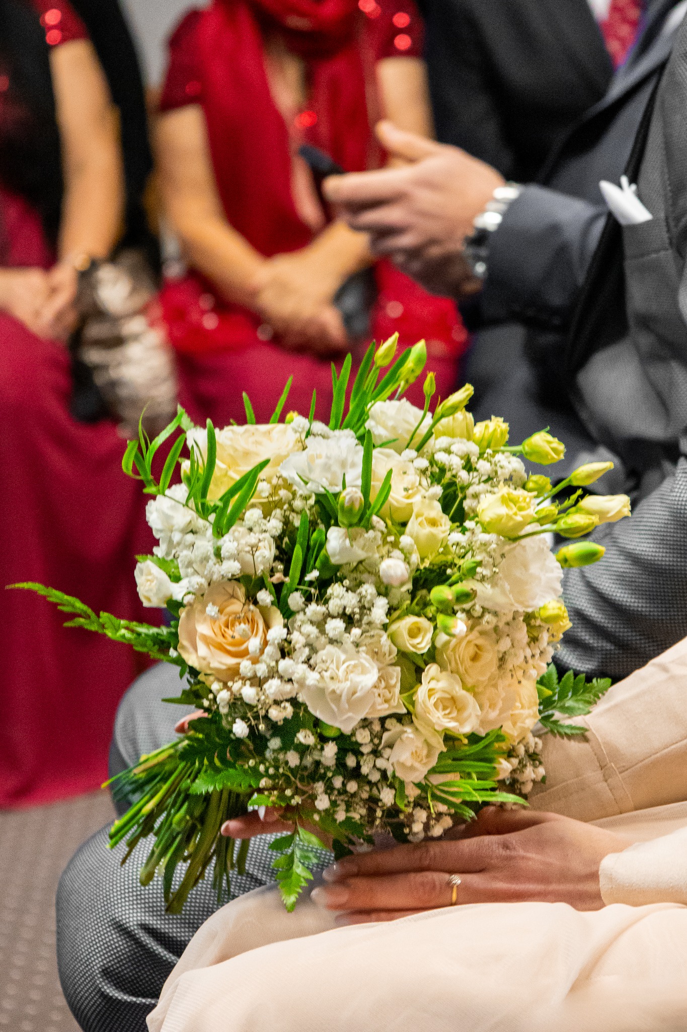 Panna młoda trzyma bukiet ślubny z kremowych róż i białych goździków na tle gości w czerwonych i szarych strojach.