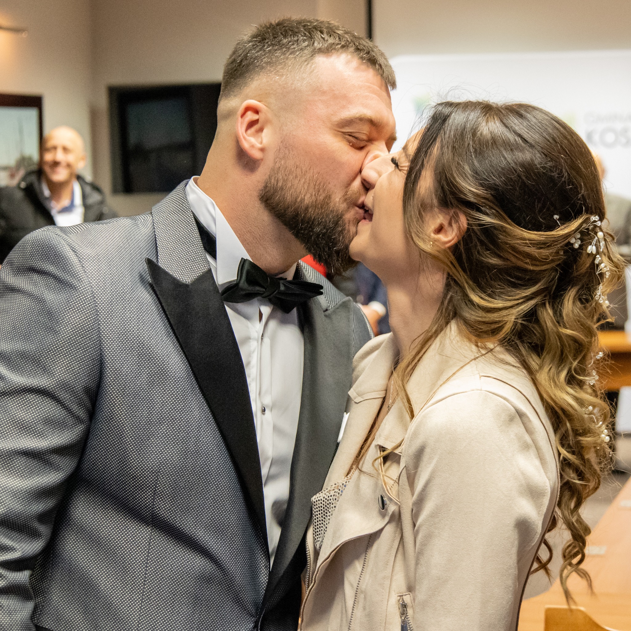 Nowożeńcy w pocałunku: mężczyzna w szarej marynarce z czarnymi klapami i muszką, kobieta w beżowej kurtce i z upiętymi włosami z ozdobami, ujęcie z bliska.