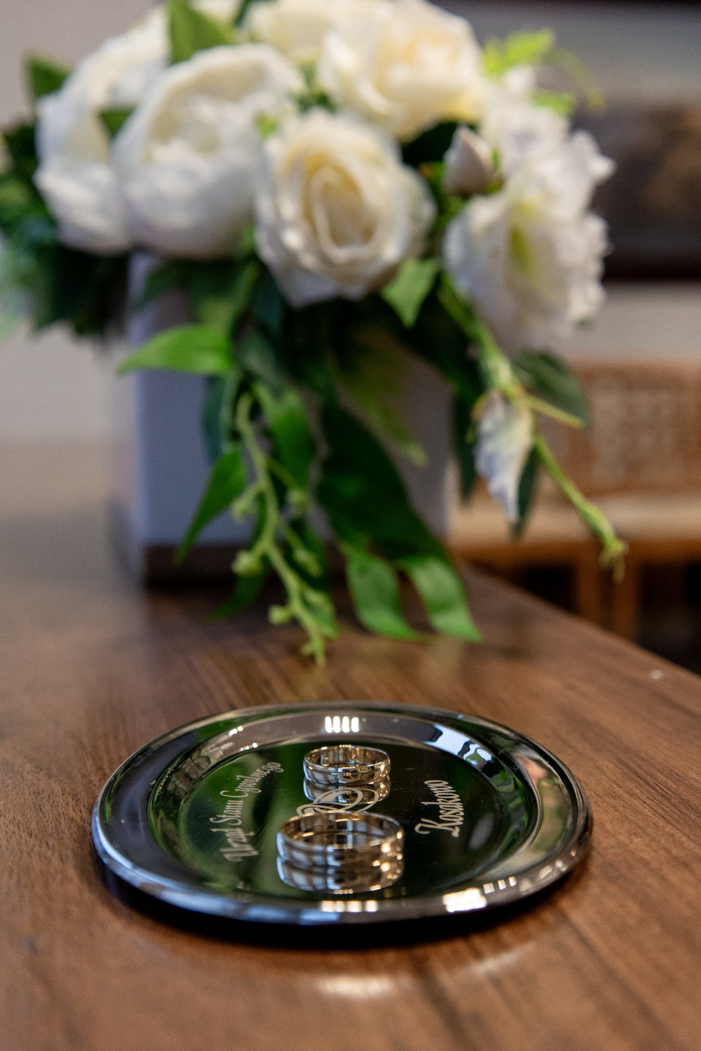 Dwa złote obrączki ślubne na srebrnej tacy z grawerem, w tle bukiet białych kwiatów i drewniany stół.