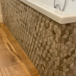 Fragment obudowy wanny z mozaiką 3D w odcieniach beżu, sąsiadującej z podłogą wyłożoną panelami imitującymi jasne drewno.