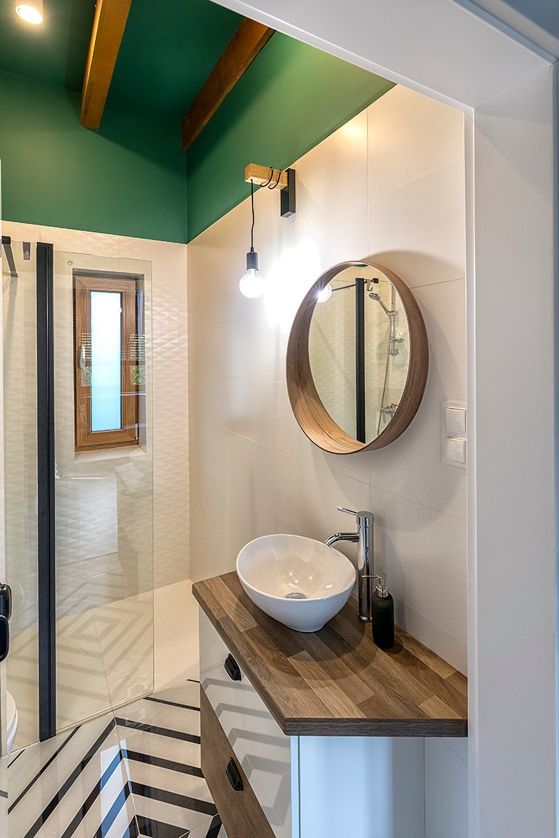 Nowoczesna łazienka z zielonym sufitem, drewnianymi belkami, okrągłym lustrem w drewnianej ramie, białą umywalką nablatową i kabiną prysznicową z czarnymi profilami.