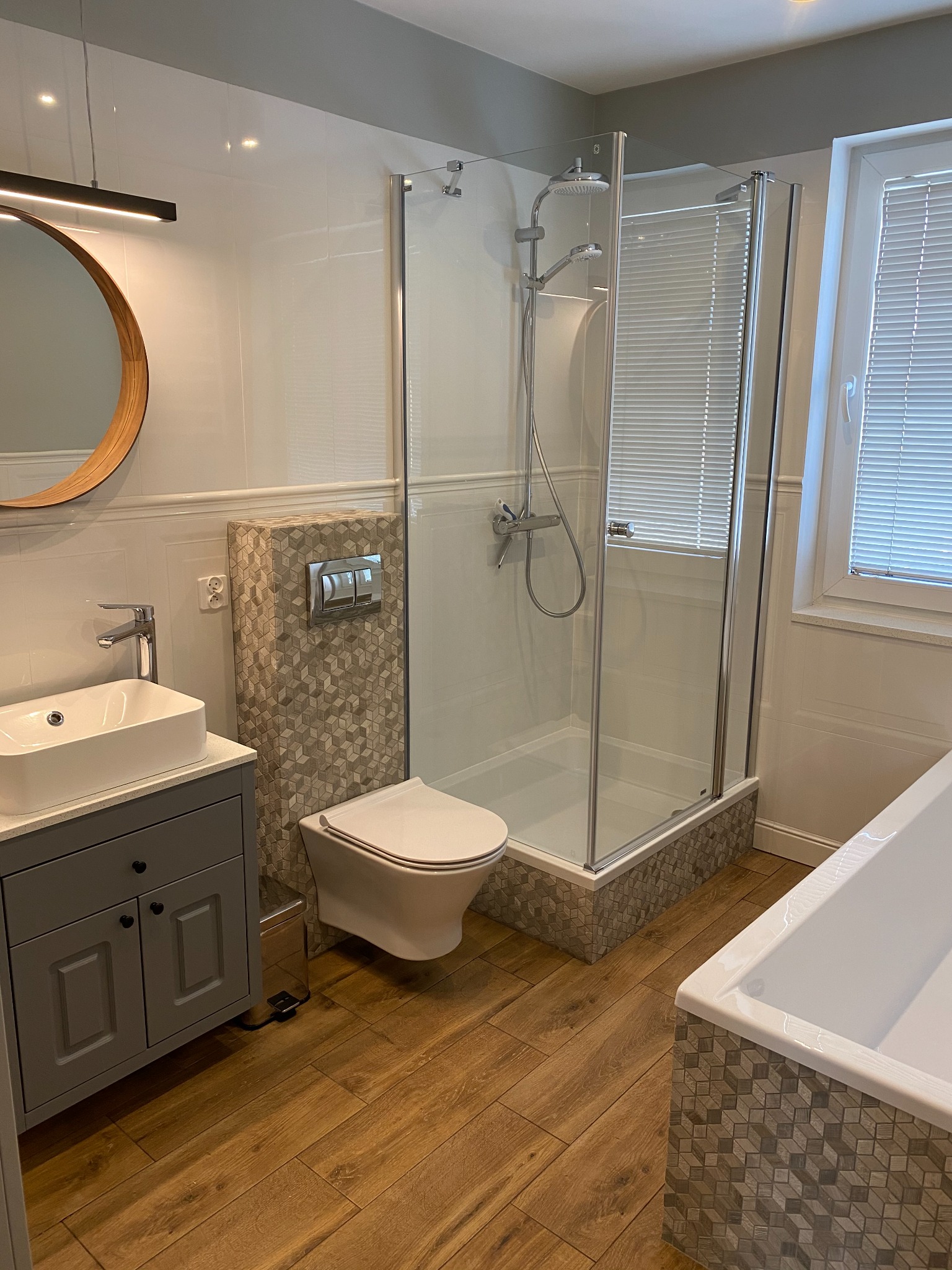 Nowoczesna łazienka z drewnopodobną podłogą, kabiną prysznicową ze szklanymi drzwiami, umywalką nablatową na szarej szafce i okrągłym lustrem z drewnianą ramą. Akcent dekoracyjny stanowi mozaika 3D...