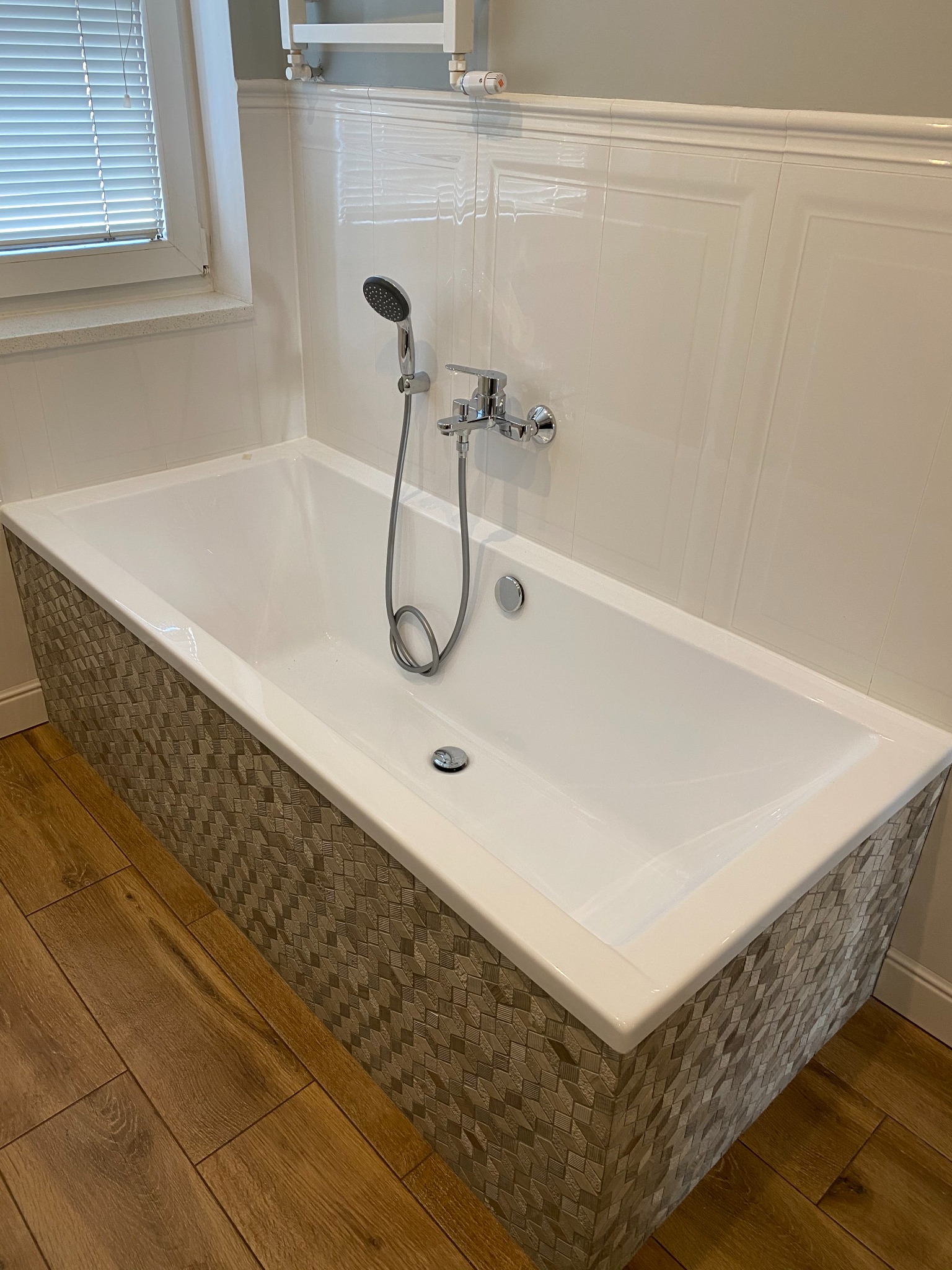 Biała wanna z mozaikową obudową, chromowana bateria z prysznicem, białe płytki na ścianie i drewnopodobna podłoga w łazience z oknem.