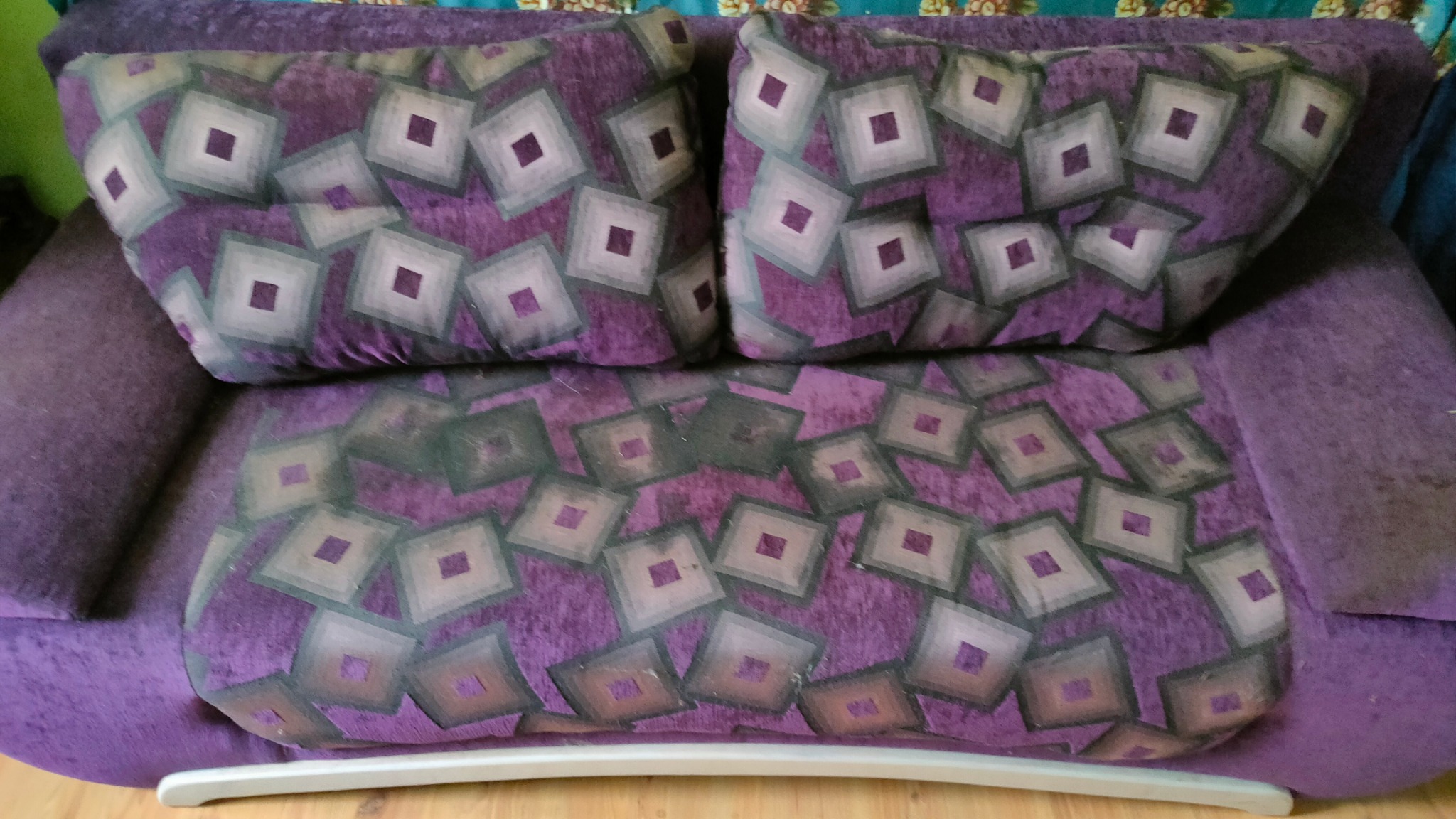 Fioletowa kanapa z poduszkami w geometryczne wzory, widoczne plamy na siedzisku.