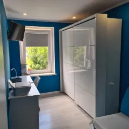 Pokój z intensywnie niebieskimi ścianami, białymi meblami (szafa przesuwna, biurko) i jasną podłogą; widoczne okno z roletą i telewizor na ścianie.